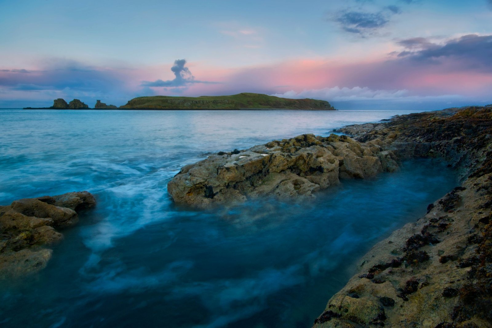 A tranquil seascape with rocky shores and an island in the distance during twilight on Northern Ireland's coast.