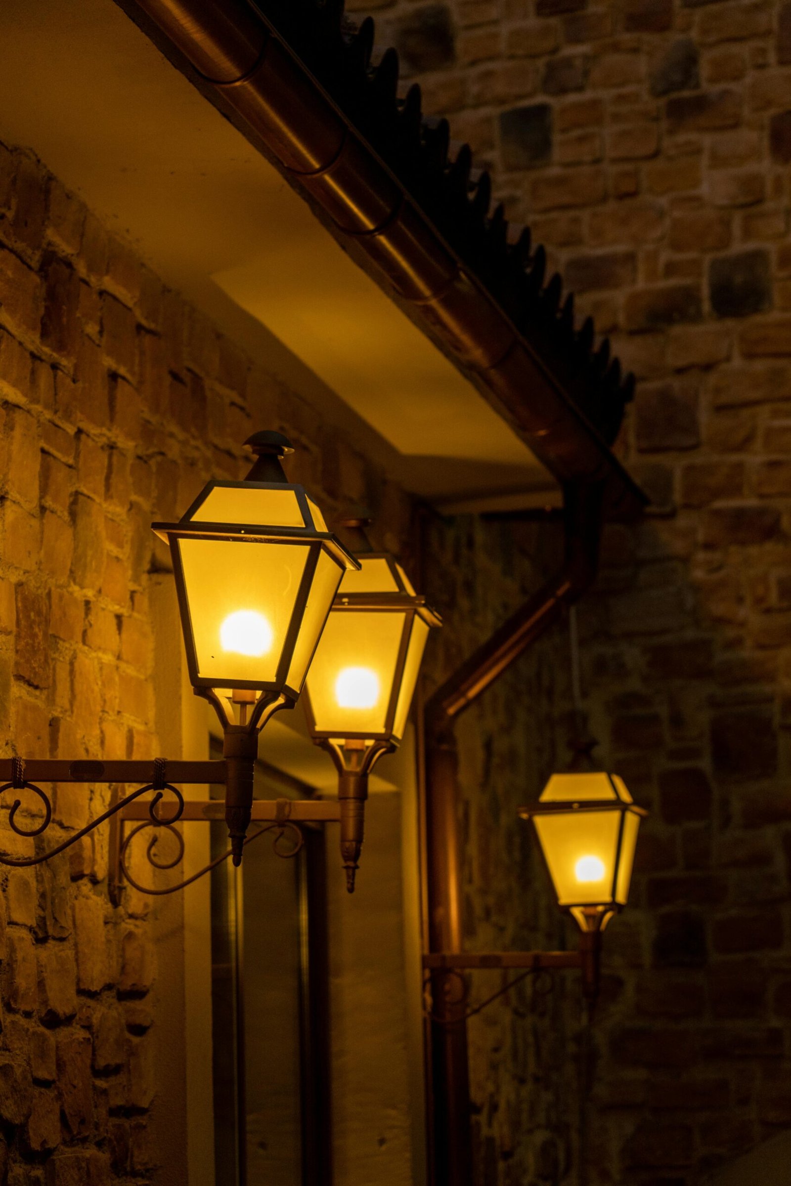Glowing street lanterns attached to a textured brick wall illuminate the evening atmosphere.